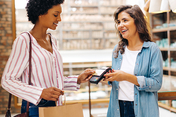 Two women stand smiling while one taps a credit card on a wireless card reader at a local business.