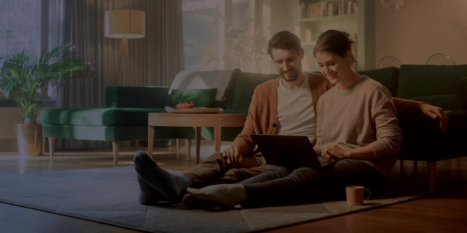 Young couple sit on the floor of their home smiling and using their internet connected devices.