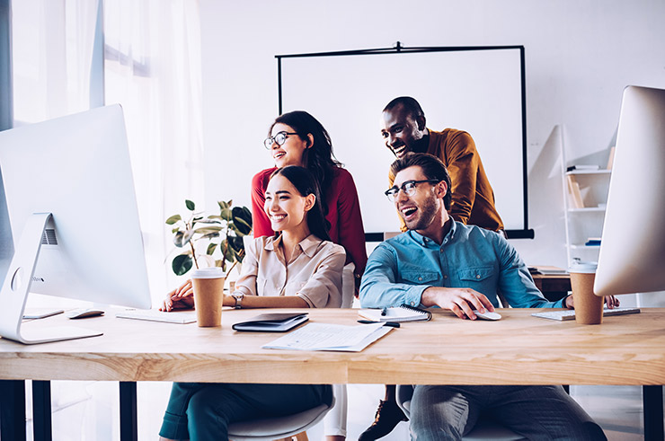 Four work colleagues sit behind a long desk with multiple computers all laughing and looking at the screens.